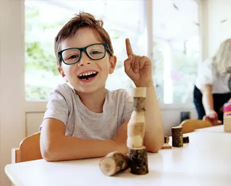 Un garçon souriant à la crèche cocon Kids joue avec des blocs de construction en bois et lève le doigt avec enthousiasme.